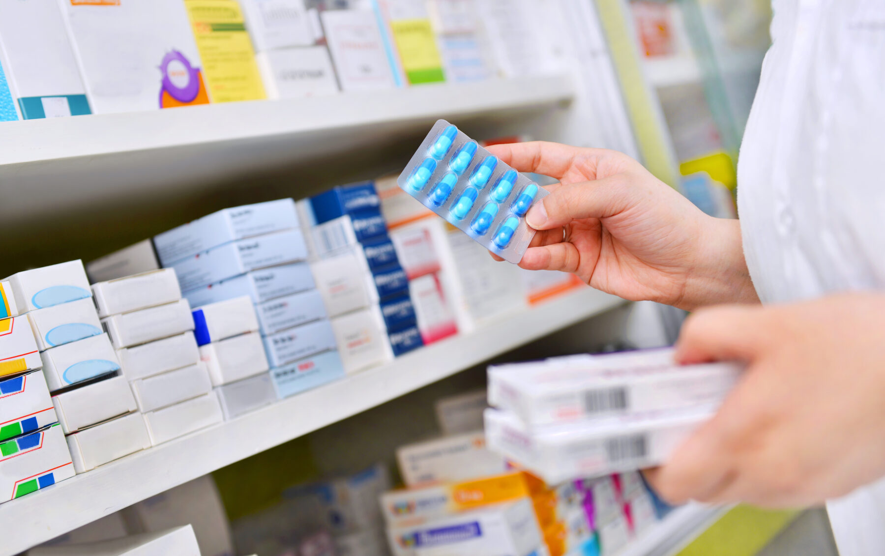 A hand holds a packet of blue capsules in front of shelves of medication boxes.