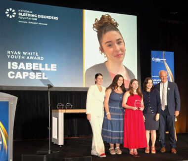 A small group of people stand on a stage around a girl in a red dress who is holding an award with her image projected on a screen behind her.