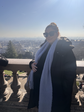 A woman wears a dark coat and sunglasses with a long purple scarf while standing in front of a stone railing overlooking a city. She is seven months pregnant and has her right hand resting on her belly.